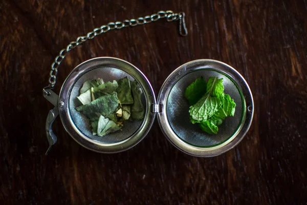 Tee-Ei aus Metall mit getrockneten Blättern von grünem Tee und frischer Minze auf dunkelbraunem Holzhintergrund — Stockfoto