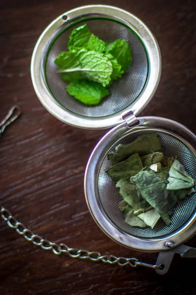 Infusor de té de metal con hojas secas de té verde y menta fresca sobre un fondo de madera marrón oscuro — Foto de Stock