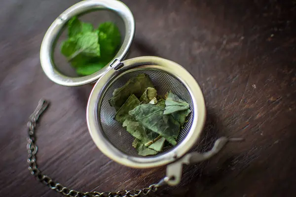 Infusor de té de metal con hojas secas de té verde y menta fresca sobre un fondo de madera marrón oscuro — Foto de Stock