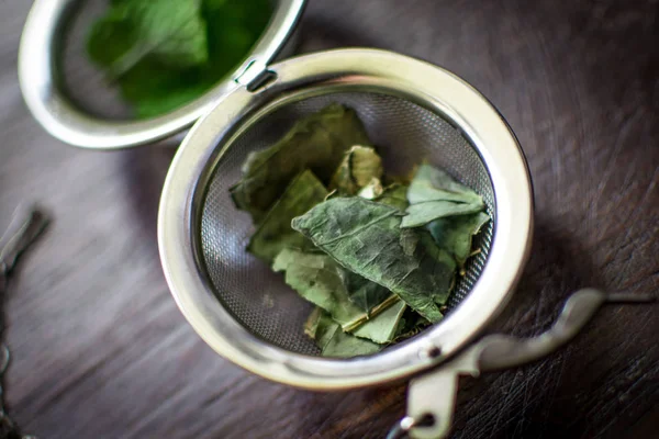 Infuseur de thé en métal avec des feuilles séchées de thé vert et de menthe fraîche sur un fond en bois brun foncé — Photo