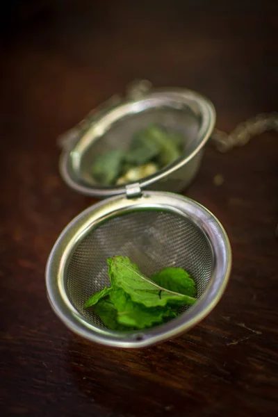 Infusor de té de metal con hojas secas de té verde y menta fresca sobre un fondo de madera marrón oscuro — Foto de Stock