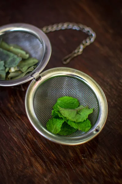 Infusor de té de metal con hojas secas de té verde y menta fresca sobre un fondo de madera marrón oscuro — Foto de Stock