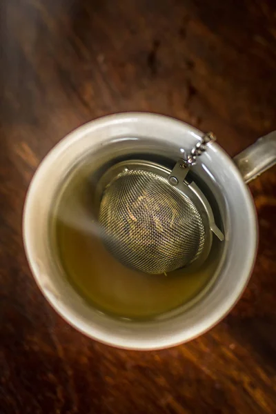 Xícara de chá com infusor em água fumegante quente com chá verde, ervas de hortelã e mel em um fundo de madeira escura — Fotografia de Stock