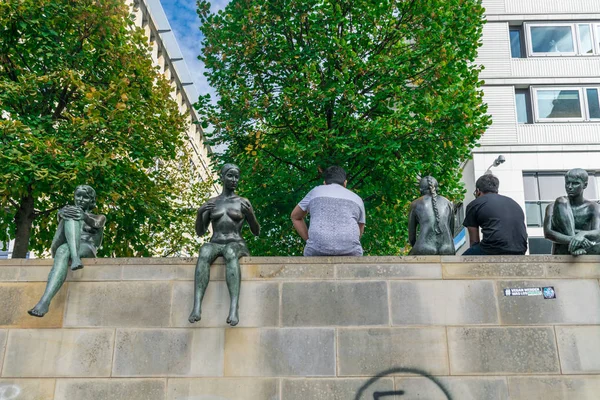 BERLIM, ALEMANHA - 26 de setembro de 2018: Visitantes sentados em uma parede entre as Três meninas e um menino um conjunto de estátuas de bronze retratando jovens e nus sentados em uma parede perto do rio Spree — Fotografia de Stock