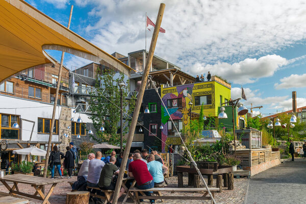 BERLIN, GERMANY - September 26, 2018: Colorful overview of a group of men sitting in wood benches at the Pampa beach bar and chilling while having a beer in the Holzmarkt, near the Spree river