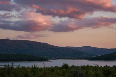Göl ve orman üzerinde pembe bulutlar. Parlak günbatımı ışığı. Mor ve kırmızı gözlükler. Gün batımında kırsal alan. Dağları ve gölü olan taşralı bir motif.