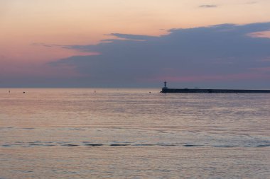 Gün batımına karşı bir deniz feneri. Güzel, huzurlu deniz manzarası. Deniz ufkunda küçük gemiler. Mor bulutlar ve yumuşak pembe su. Liman. Gün batımında martılar. Atmosferik akşam manzarası.