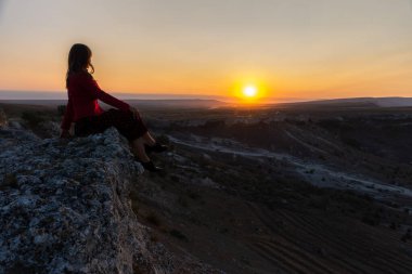 Kız uçurumun kenarında oturur ve güneşe bakar. Koyu renk saçlı güzel bir kız. Dağın arka planında elbise giymiş poz veriyor. Dağlarda şafak vakti. Doğayla baş başa bir kız..