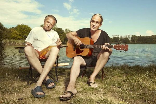Two older men relaxing and singing with guitar by summer lake