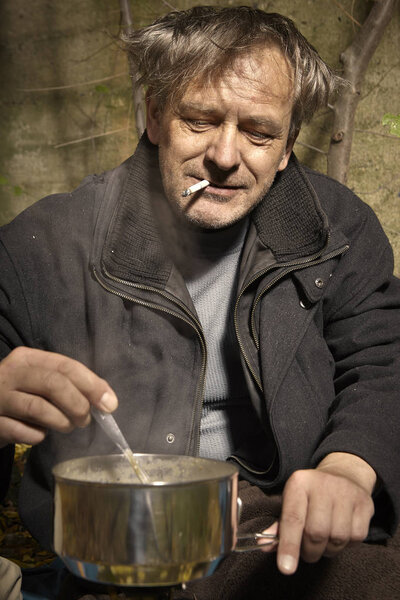 Ugly pauper man living outdoor preparing and eating lentil soup