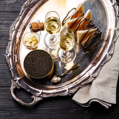 Black caviar in can, fresh bread toast and champagne on silver tray on black wooden background