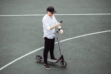 Modern insan Street elektrikli scooter ile duran onun smartphone kullanarak şık kıyafeti