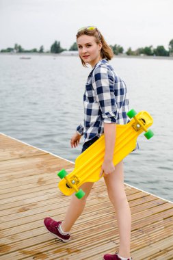 Sarı longboard elinde tutan ve ahşap bir iskele üzerinde yürüme hipster kıyafeti genç neşeli kız