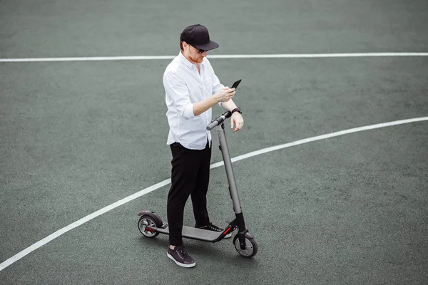 Pria modern dalam pakaian bergaya menggunakan smartphone sambil berdiri di jalan dengan skuter listrik — Stok Foto