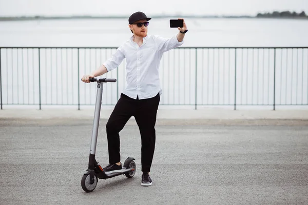 Сучасний чоловік у стильному вбранні робить селфі, стоячи на вулиці з електричним скутером — стокове фото