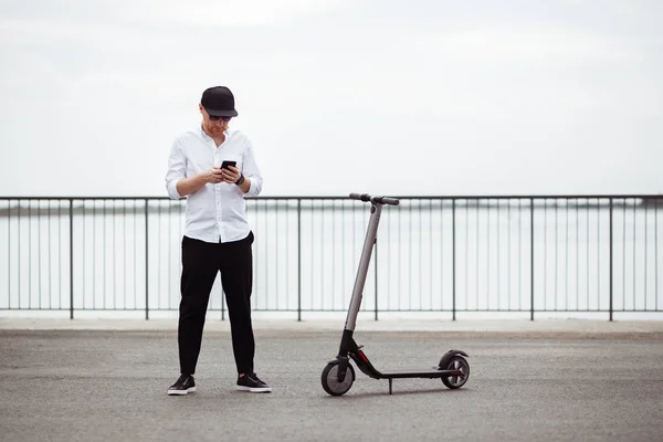 Homem moderno em roupa elegante usando seu smartphone enquanto está de pé na rua com scooter elétrico — Fotografia de Stock