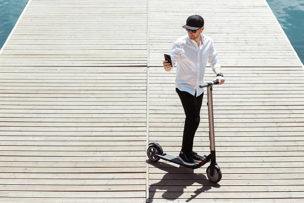 Moderne mens gekleed wit overhemd en een zwarte broek met behulp van zijn telefoon terwijl staande op een houten pier met elektrische scooter — Stockfoto