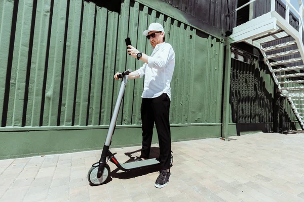 Hombre moderno en traje elegante usando su teléfono inteligente mientras está de pie en la calle con scooter eléctrico — Foto de Stock
