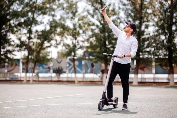 Pria modern dalam pakaian stylish membuat selfie sambil berdiri di jalan dengan skuter listrik — Stok Foto