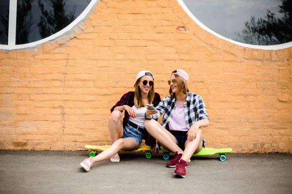 Dos jóvenes amigas felices en traje hipster sentadas en longboards y haciendo selfie en el teléfono —  Fotos de Stock