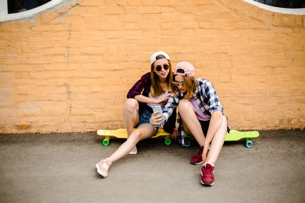 Due giovani amici felici ragazza in abito hipster seduti su longboard e fare selfie al telefono — Foto Stock