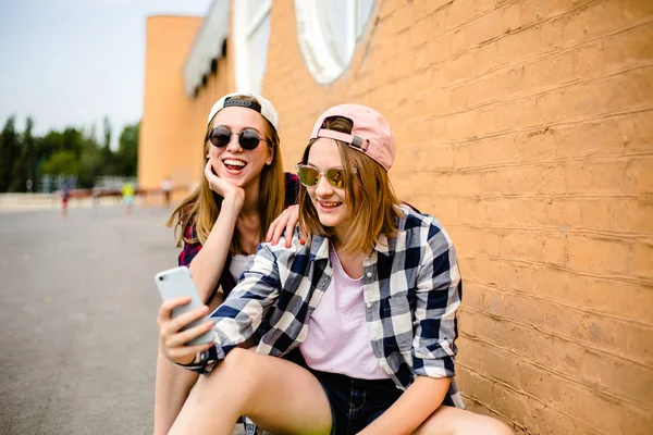 Due giovani amici felici ragazza in abito hipster seduti su longboard e fare selfie al telefono — Foto Stock