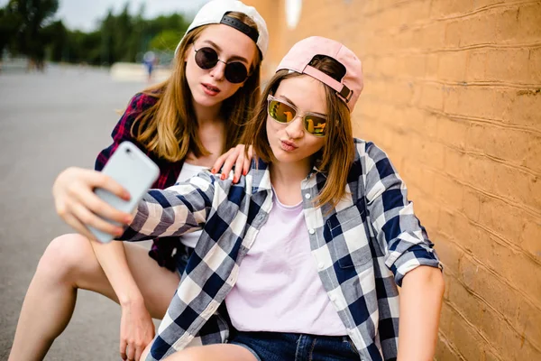 Två unga glada väninnor i hipster outfit sitter på longboards och göra selfie på telefon — Stockfoto