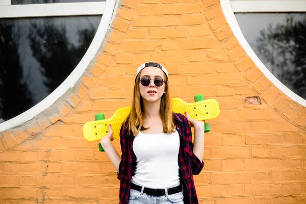 Молода весела дівчина позує з жовтим скейтбордом на помаранчевій стіні — стокове фото