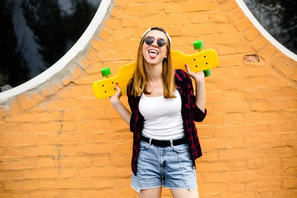 Jovem menina alegre posando com skate amarelo contra a parede laranja — Fotografia de Stock