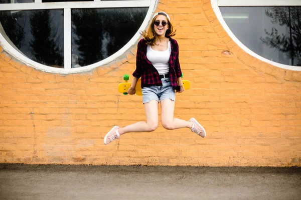 Jovem menina alegre posando com skate amarelo contra a parede laranja — Fotografia de Stock