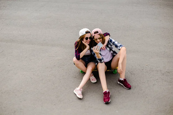 Duas jovens amigas felizes em roupa hipster sentadas juntas no longboard e fazendo selfie no telefone — Fotografia de Stock