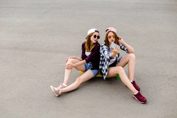 Two young happy girl friends in hipster outfit sitting together on longboard and making selfie on phone — Stock Photo, Image