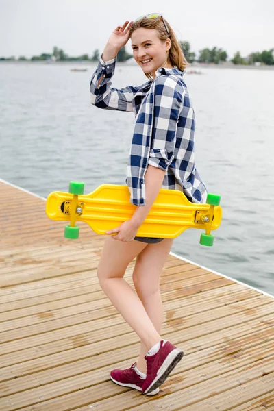 Jovem menina alegre em roupa hipster segurando longboard amarelo em sua mão e andando em um cais de madeira — Fotografia de Stock