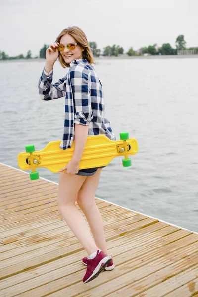 Fiatal vidám lány csípő felszerelés sárga longboard tartja a kezében, és egy fából készült móló séta — Stock Fotó