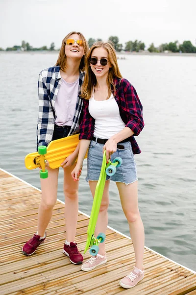 Duas alegres meninas skatistas felizes em roupa hipster se divertindo em um cais de madeira durante as férias de verão — Fotografia de Stock