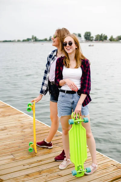 Dos alegres chicas patinadoras felices en traje hipster divirtiéndose en un muelle de madera durante las vacaciones de verano —  Fotos de Stock