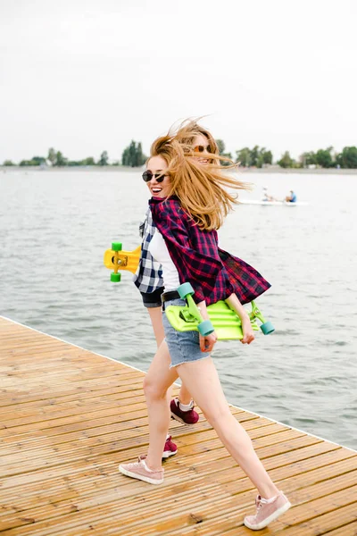 Dos alegres chicas patinadoras felices en traje hipster divirtiéndose en un muelle de madera durante las vacaciones de verano —  Fotos de Stock