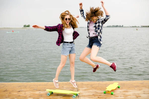 Dos alegres chicas patinadoras felices en traje hipster divirtiéndose en un muelle de madera durante las vacaciones de verano —  Fotos de Stock