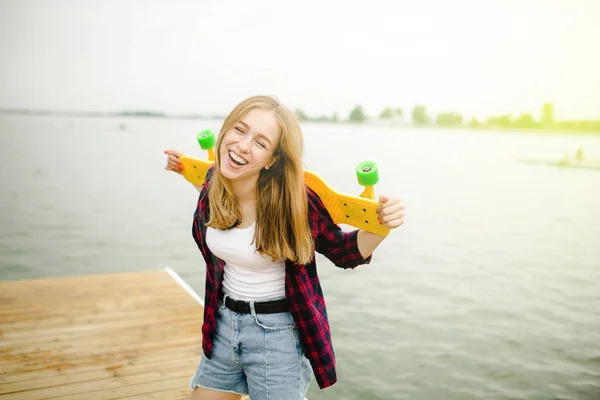 Alegre feliz skatista menina em roupa hipster se divertindo em um cais de madeira durante as férias de verão — Fotografia de Stock