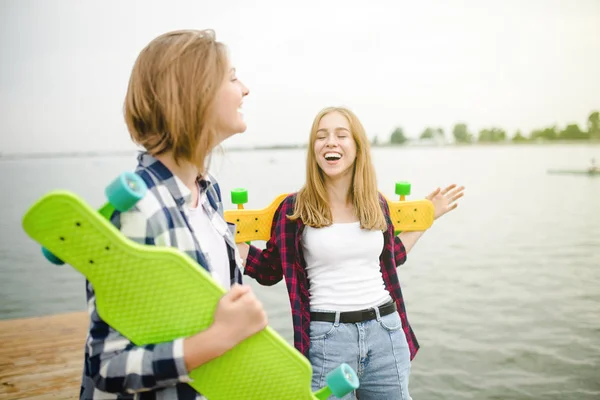 Dos alegres chicas patinadoras felices en traje hipster divirtiéndose en un muelle de madera durante las vacaciones de verano —  Fotos de Stock