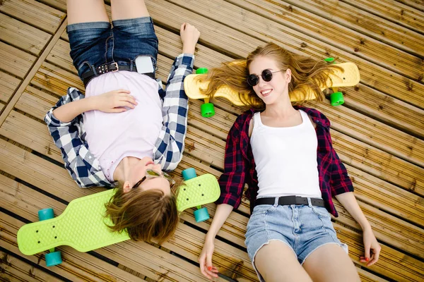 Twee stijlvolle jonge stedelijke meisjes met longboards liggen op de houten vloer in de straat. Vrienden hebben plezier en tijd doorbrengen samen in de zomer tijdens de vakantie — Stockfoto