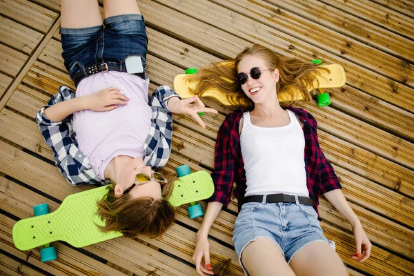 Twee stijlvolle jonge stedelijke meisjes met longboards liggen op de houten vloer in de straat. Vrienden hebben plezier en tijd doorbrengen samen in de zomer tijdens de vakantie — Stockfoto