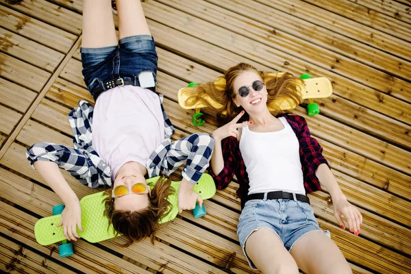 Twee stijlvolle jonge stedelijke meisjes met longboards liggen op de houten vloer in de straat. Vrienden hebben plezier en tijd doorbrengen samen in de zomer tijdens de vakantie — Stockfoto