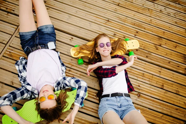 Δύο όμορφα νεαρά κορίτσια αστικών με longboards βρίσκονται στο ξύλινο δάπεδο στο δρόμο. Φίλους να διασκεδάσουν και να περάσουν χρόνο μαζί το καλοκαίρι κατά τη διάρκεια των διακοπών — Φωτογραφία Αρχείου