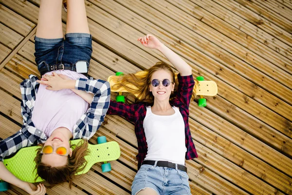Dos chicas urbanas jóvenes con estilo con longboards se encuentran en el suelo de madera en la calle. Los amigos se divierten y pasan tiempo juntos en verano durante las vacaciones. —  Fotos de Stock