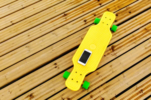 Telefon ahşap zemin üzerinde yer alan bir sarı plastik longboard üzerinde yatıyor. Üstten görünüm — Stok fotoğraf