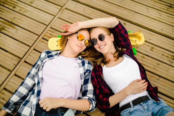 Dos chicas urbanas jóvenes con estilo con longboards se encuentran en el suelo de madera en la calle. Los amigos se divierten y pasan tiempo juntos en verano durante las vacaciones. —  Fotos de Stock