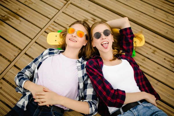 Due giovani ragazze alla moda urbane con longboard si trovano sul pavimento in legno in strada. Gli amici si divertono e trascorrono del tempo insieme in estate durante le vacanze — Foto Stock