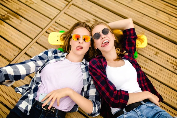 Två snygga unga urbana flickor med longboards ligga på trä golv i gatan. Vänner ha kul och umgås tillsammans på sommaren under semestern — Stockfoto