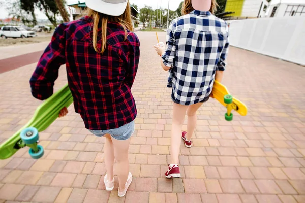 Duas meninas urbanas com longboards vestindo camisas quadriculadas que vão ao longo da rua na cidade — Fotografia de Stock